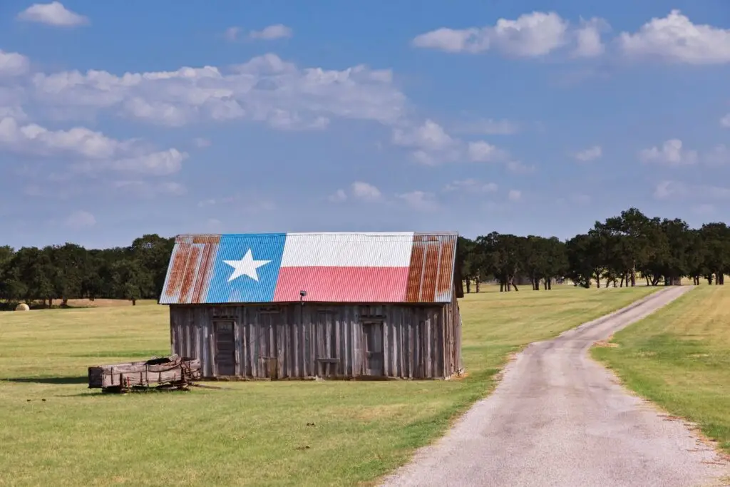 Rustic barn with Texas flag roof in open field, surrounded by trees and gravel road, representing local culture in Katy, Texas.